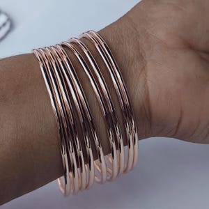 以下が含まれることがあります： 手首に重ね付けされたローズゴールドのバングル。バングルは滑らかで丸く、光を反射しています。ジュエリーは温かみのあるメタリックカラーで、クラシックでエレガントなスタイルを表現しています。
