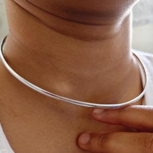 Könnte beinhalten: Eine silberfarbene Choker-Halskette. Die Halskette ist ein einfaches, dünnes und abgerundetes Band, das eng am Hals anliegt. Das Bild zeigt die Halskette beim Tragen, mit einer Hand, die sie sanft berührt.