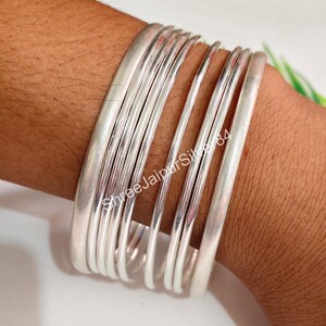 Könnte beinhalten: Eine Ansammlung silberner Armreifen unterschiedlicher Dicke ist auf einem Handgelenk gestapelt. Die Armreifen haben eine matte Oberfläche und reflektieren das Licht. Der Text "Shree Jaipur Silver 84" ist im Bild zu sehen. Diese Armreifen sind eine Art Schmuck.