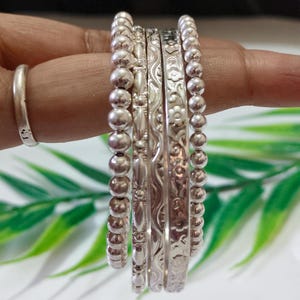 Könnte beinhalten: Eine Sammlung von Silberringen und Armreifen. Die Ringe umfassen ein schlichtes Band, während die Armreifen verschiedene Designs aufweisen, darunter Perlen- und Blumenmuster. Der Schmuck wird auf einem Finger präsentiert, wobei die verschiedenen Texturen und Stile hervorgehoben werden.