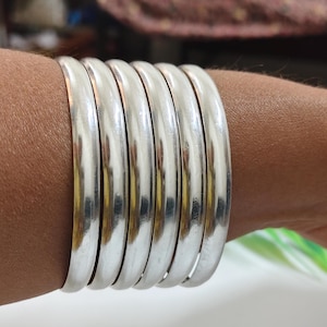 Puede incluir: Una colección de siete brazaletes plateados, cada uno con un diseño liso y redondeado. Los brazaletes están apilados en una muñeca, reflejando la luz y creando una apariencia metálica brillante. Joyería sencilla y elegante.