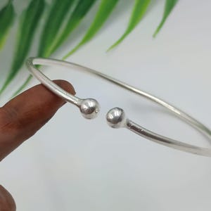 Puede incluir: Un brazalete abierto de color plateado con un diseño liso y redondeado. El brazalete presenta un estilo sencillo y elegante con una pequeña bola en cada extremo abierto. La joya se exhibe sobre un fondo blanco con hojas verdes.