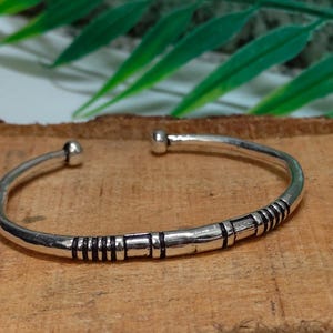 K&ouml;nnte beinhalten: Ein silberfarbener Armreif aus Metall mit einem glatten, abgerundeten Band und einer kleinen kugelf&ouml;rmigen Verzierung an jedem Ende. Die Oberfl&auml;che des Armreifs ist mit einer Reihe von dunklen, vertieften horizontalen Linien und B&auml;ndern verziert.