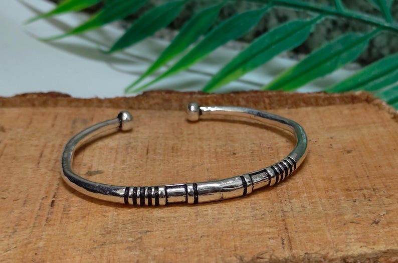 K&ouml;nnte beinhalten: Ein silberfarbener Armreif aus Metall mit einem glatten, abgerundeten Band und einer kleinen kugelf&ouml;rmigen Verzierung an jedem Ende. Die Oberfl&auml;che des Armreifs ist mit einer Reihe von dunklen, vertieften horizontalen Linien und B&auml;ndern verziert.