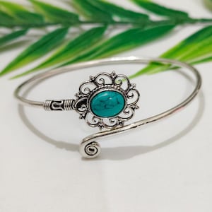 Könnte beinhalten: Ein silberfarbenes Armband in offener Form mit einem zentralen ovalen Türkisstein, der in einen filigranen floralen Rahmen gefasst ist. Das Metallband hat eine polierte Oberfläche mit einem Spiralabschluss und einem strukturierten Detail.