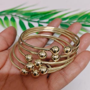 Könnte beinhalten: Ein Satz goldfarbener Armreifen mit kugelförmigen Akzenten. Die Armreifen sind gestapelt und auf einer Hand präsentiert, was ihre glänzende, metallische Oberfläche hervorhebt. Das Design umfasst mehrere dünne Bänder, die durch die kugelförmigen Verzierungen verbunden sind.