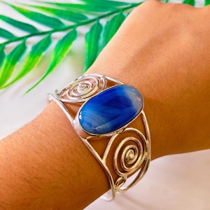 May include: Silver cuff bracelet with a large, oval, blue gemstone. The bracelet has an openwork design with spiral and swirl details. A decorative piece of jewellery.