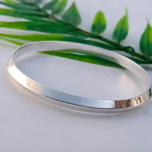 Puede incluir: Un brazalete rígido de tono plateado pulido con un diseño de borde biselado liso. La banda circular de metal presenta una superficie reflectante que capta la luz, creando una estética limpia y sencilla para este accesorio de muñeca.