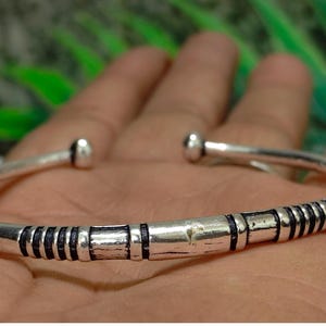 Könnte beinhalten: Ein silberfarbenes, offenes Armband mit polierter Oberfläche, das in der Mitte dunkle, vertiefte Rillen aufweist und an beiden Enden mit abgerundeten Kugelabschlüssen versehen ist, was einen deutlichen Kontrast zwischen Metall und Linien erzeugt.