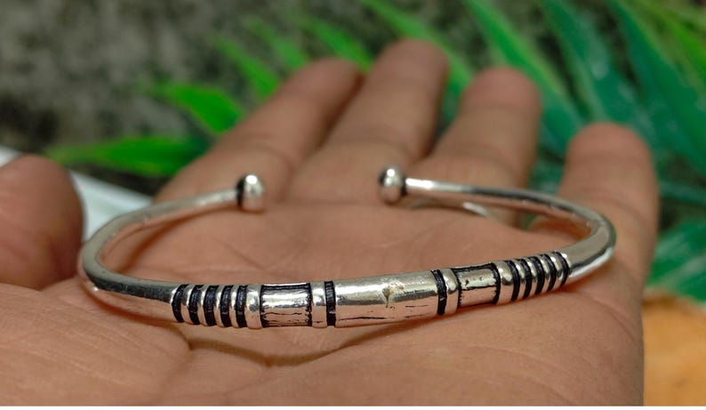 K&ouml;nnte beinhalten: Ein silberfarbenes, offenes Armband mit polierter Oberfl&auml;che, das in der Mitte dunkle, vertiefte Rillen aufweist und an beiden Enden mit abgerundeten Kugelabschl&uuml;ssen versehen ist, was einen deutlichen Kontrast zwischen Metall und Linien erzeugt.