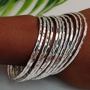 Könnte beinhalten: Eine Sammlung silberner, gehammerter Armreifen. Die Armbänder sind auf einem Arm gestapelt und zeigen ihre strukturierte Oberfläche und reflektierende Qualität. Die Armreifen sind hellsilberfarben.