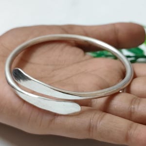 Può includere: Un bracciale a polsino argentato con un design curvo e aperto. Il bracciale presenta due estremità allungate a forma di goccia che si sovrappongono leggermente. Il metallo liscio e lucido riflette la luce, creando un'estetica minimalista.