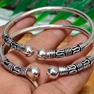 Puede incluir: Dos pulseras plateadas con detalles decorativos en negro y plata. Las pulseras tienen un diseño abierto con extremos redondeados. Las secciones decorativas presentan un patrón intrincado. Las joyas se exhiben en una mano.