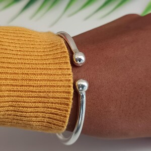 Könnte beinhalten: Ein silbernes Armreif-Armband mit abgerundeten Kugelenden. Das offene Design ermöglicht ein einfaches Tragen. Das Armband wird an einem Arm gezeigt, neben einem senfgelben Pullover.