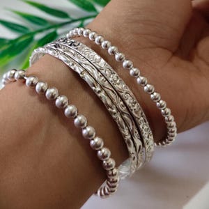 Könnte beinhalten: Eine Sammlung silberner Armbänder schmückt ein Handgelenk. Das Set umfasst Perlenarmbänder und aufwendig gestaltete Armreifen mit floralen Mustern. Die Armbänder bestehen aus einem glänzenden, metallischen Material, das Licht reflektiert. Die Gesamtästhetik ist elegant und dekorativ.