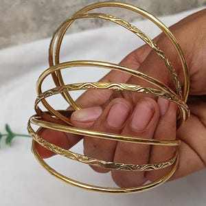May include: A collection of gold-coloured bangles. Some bangles have a simple, smooth design, while others feature an etched pattern. The bangles are stacked together, creating a layered look. The jewellery is displayed against a neutral background.