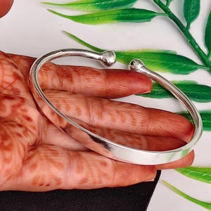 Könnte beinhalten: Ein polierter Armreif in Silberoptik mit einem glatten, offenen Design und zwei abgerundeten Kugelenden an beiden Seiten. Die Metalloberfläche ist reflektierend und schlicht gehalten, was eine einfache, kreisförmige Form ergibt, die leicht anpassbar ist.