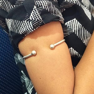 Könnte beinhalten: Ein Oberarmreif aus silberfarbenem Metall mit offenem Design und einer polierten Kugel an jedem Ende. Das Armband wird am Oberarm getragen und zeichnet sich durch eine glatte, reflektierende Metalloberfläche und eine schlichte Formgebung aus.