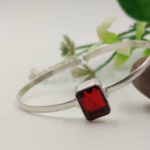 Könnte beinhalten: Ein silbernes Armreif-Armband mit einem rechteckigen, facettierten, tiefroten Edelstein. Das Armband hat ein schlichtes, elegantes Design mit poliertem Finish. Der Edelstein ist der Mittelpunkt, gefasst in einer quadratischen Silberfassung. Das Armband wird auf einer weißen Oberfläche präsentiert.