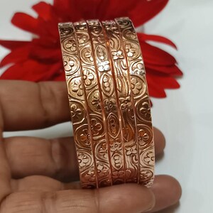 Könnte beinhalten: Sechs kupferfarbene Armreifen mit geprägtem Blumenmuster. Die Armreifen werden in einer Hand gehalten, mit einer roten Blume im Hintergrund. Die Armreifen haben eine Breite von etwa 1,3 cm.