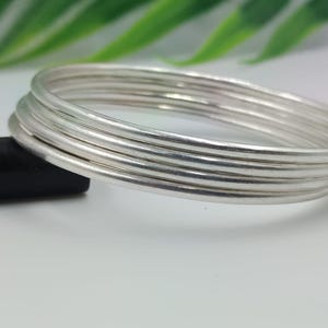 Könnte beinhalten: Ein Stapel von sechs silbernen Armreifen. Die Armbänder sind rund und glatt, mit einem glänzenden, metallischen Finish. Die Armreifen sind übereinander gestapelt, wodurch ein geschichteter Effekt entsteht. Der Hintergrund ist verschwommen, mit grünen Blättern und einer weißen Oberfläche.