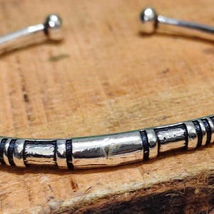 K&ouml;nnte beinhalten: Ein silberfarbenes Armband in offener Form mit abgerundeten Kugelenden. Das Band ist mit einer Reihe von vertieften schwarzen Rillen und erhabenen silbernen Segmenten verziert, die ein sich wiederholendes Muster auf der Vorderseite bilden.