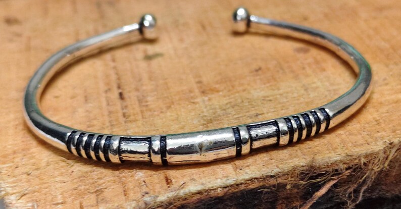 K&ouml;nnte beinhalten: Ein silberfarbenes Armband in offener Form mit abgerundeten Kugelenden. Das Band ist mit einer Reihe von vertieften schwarzen Rillen und erhabenen silbernen Segmenten verziert, die ein sich wiederholendes Muster auf der Vorderseite bilden.
