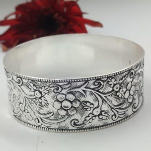 Könnte beinhalten: Ein silberfarbenes Armreif-Armband mit einem eingravierten Blumenmuster. Das Armband hat ein breites Band mit detaillierten Schnitzereien von Blumen und Blättern. Eine rote Blume ist im Hintergrund.