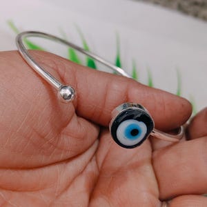 Könnte beinhalten: Ein silberfarbenes Armband mit offenem Design. Es hat eine kleine silberne Kugel an einem Ende und einen kreisförmigen Anhänger mit einem blauen Augendesign am anderen. Der Augenanhänger hat einen schwarzen Hintergrund mit einem weißen und blauen Auge.