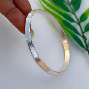 Könnte beinhalten: Ein silberfarbenes Armreif-Armband mit glatter, polierter Oberfläche. Das Armband ist rund und hat ein schlichtes, elegantes Design. Das Bild enthält auch eine grüne Pflanze und eine Hand.