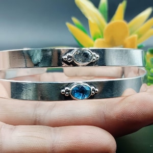 Könnte beinhalten: Zwei silberne Armreifen, jeweils mit einem ovalen Edelstein. Ein Armreif hat einen klaren Edelstein, der andere einen leuchtend blauen Stein. Die Armreifen werden vor einem verschwommenen gelb-grünen Hintergrund präsentiert.