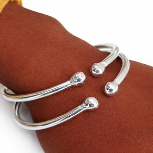 May include: A silver cuff bracelet with a rounded, open design. The bracelet has two parallel bands that curve around the wrist, each ending in a small, spherical accent. The jewellery is displayed on a brown skin tone.