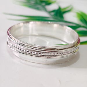 Könnte beinhalten: Ein polierter Silberarmreif mit einem Mittelband aus feiner Perlenstruktur, das von zwei glatten, schlichten Silberbändern flankiert wird. Das Metall hat eine helle, reflektierende Oberfläche, die ein klares und klassisches Erscheinungsbild erzeugt.