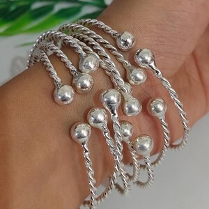 Può includere: Un insieme di braccialetti argentati. Ogni braccialetto presenta un design a corda intrecciata e una piccola sfera rotonda all'estremità di un'estensione curva. I braccialetti sono sovrapposti.