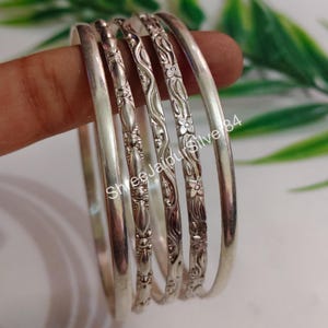 Könnte beinhalten: Eine Reihe von Silberarmreifen, einige mit floralen und gemusterten Designs. Die Armreifen werden vor einem weichen Hintergrund mit grünem Laub gezeigt. Der Text "Shree Jaipur Silver 84" ist sichtbar.