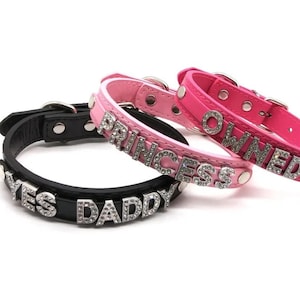 May include: Three pet collars in black and pink leather. The black collar has the text "YES DADDY" in silver rhinestones. The pink collars have the text "PRINCESS" and "OWNED" in silver rhinestones. Each collar has a silver buckle.