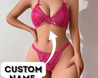 May include: A hot pink lingerie set featuring a halter-style bra with the text "GOOD GIRL" and a matching thong. The bra and thong are adorned with delicate lace detailing. The image also includes a white arrow and the text "CUSTOM NAME".