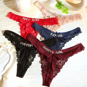 Può includere: Una collezione di perizomi in pizzo in vari colori, tra cui rosso, nero, blu navy e bordeaux. Ogni perizoma presenta testo con strass come "SEXY BABY", "EAT ME", "DADDY'S GIRL" e "CUSTOM". La biancheria intima ha un delicato design floreale in pizzo.