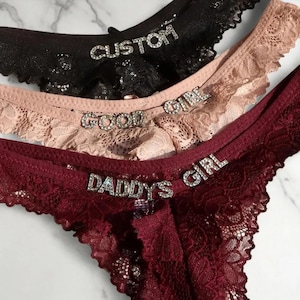 May include: Close-up of three pairs of lace underwear in black, nude, and burgundy. Each pair has rhinestone embellishments spelling out "CUSTOM", "GOOD GIRL", and "DADDY'S GIRL". The lace has a scalloped edge.
