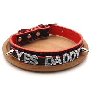 Pode incluir: Uma coleira de couro preta e vermelha com o texto "YES DADDY" em letras de strass prateadas. A coleira tem pontas prateadas e uma fivela prateada.