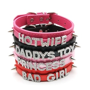 May include: A stack of leather collars in pink, black, and red, each with silver text spelling out "HOTWIFE", "DADDYS TOY", "PRINCESS", and "BAD GIRL". The collars have silver spikes and buckles.