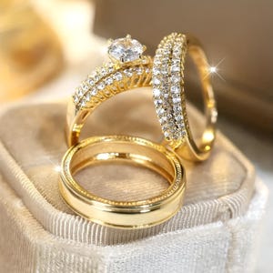 Peut inclure: Trois bagues en or, dont une avec une grosse pierre précieuse transparente et de petites pierres, et deux anneaux incrustés de petites pierres transparentes. Les bagues sont présentées sur une surface douce et claire, créant une sensation de luxe. Les bagues sont probablement pour un mariage ou des fiançailles.