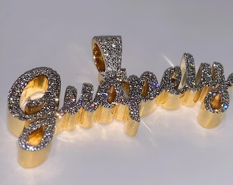 May include: A gold pendant spelling out "Guapely" in a cursive font. The letters are outlined in gold and encrusted with small, sparkling stones. The pendant has a loop at the top for a chain.