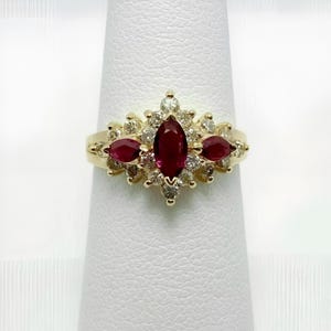 May include: A gold ring featuring a central marquise-cut ruby, flanked by two smaller rubies, and surrounded by a cluster of small diamonds. The ring is set on a gold band, showcasing a classic and elegant design.