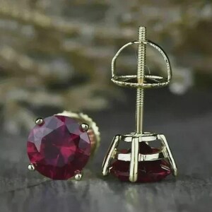 May include: A pair of gold-colored stud earrings, each featuring a large, round, deep red gemstone. The earrings have a classic design with a screw-back post for secure wear. The gemstones are faceted to enhance their brilliance.