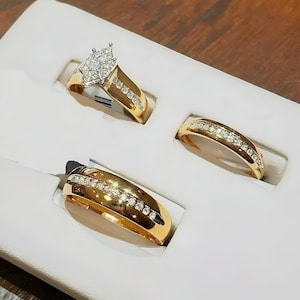May include: Three gold rings with diamond accents displayed in a white box. One ring features a cluster of diamonds in a marquise shape. The other two rings have a band of diamonds set in gold.