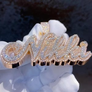 May include: A rose gold pendant spelling out the name "Nelli" in a cursive font. The pendant is encrusted with numerous small, sparkling white gemstones. The design features a layered effect, with the gemstones set in a raised pattern.
