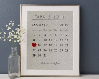 Puede incluir: Una impresión enmarcada con un calendario para enero de 2012, con el número 15 resaltado en rojo con un corazón. La impresión está sobre un fondo de algodón blanco roto texturizado e incluye los nombres "Tara & John" y la ubicación "Ashbourne, Derbyshire".