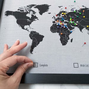 May include: A black and white world map with colourful push pins marking locations. The map is framed in black and has a grey linen background. The text "Our Travels" is printed at the top of the map. There are two checkboxes at the bottom of the map, one labelled "Complete" and the other labelled "Wish List".