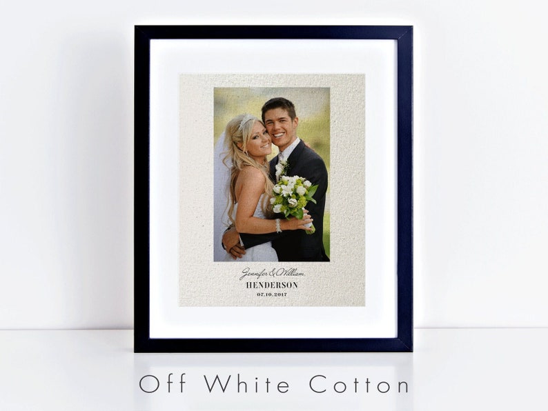 May include: A framed wedding photograph of a couple in a black frame. The photograph is printed on off-white cotton paper. The couple are smiling and the bride is holding a bouquet of white flowers. The text "Jennifer & William Henderson 07.10.2017" is printed below the photograph.